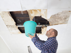 A water-damaged ceiling. A man holds a bucket to the ceiling to catch falling water.