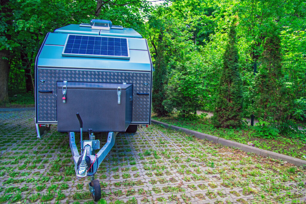 Solar panel is fixed on the tourist trailer. Off-road trailer stands in the parking lot on the background of thick green foliage.