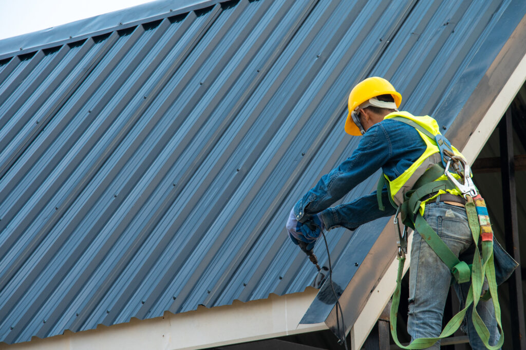 Construction worker wearing safety harness belt during working on roof structure of building on construction site,Roofer using air or pneumatic nail gun and installing metal roof tile on top new roof.